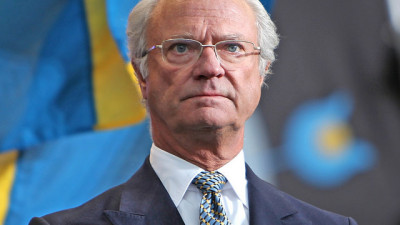 Kungen står framför några svenska flaggor. Han ser allvarlig ut och tittar ut i fjärran. Han har glest vitt hår, glasögon och en kostym med slips.