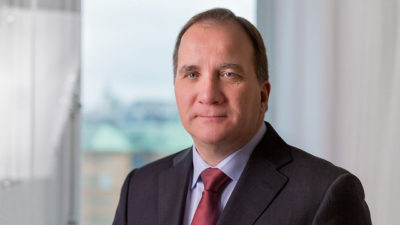 Stefan Löfven sitter i ett rum. Han har kavaj och slips på sig. Han har tunt, kort brunt hår och ganska buskiga ögonbryn. Han har stor näsa och små ögon. Han tittar in i kameran och ser ganska allvarlig ut.