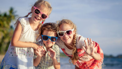 Tre syskon i solglasögon med olika färger ler framför havet.