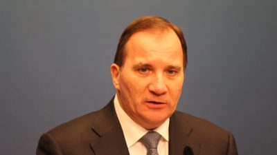 Stefan Löfven står och pratar med en mikrofon framför sig. Han har kavaj, skjorta och slips. Han har tunt brunt hår, små ögon och en stor näsa.