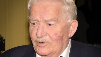 Hasse Alfredson. Han är ganska gammal och har vitt hår. Han har kostym och fluga på sig.