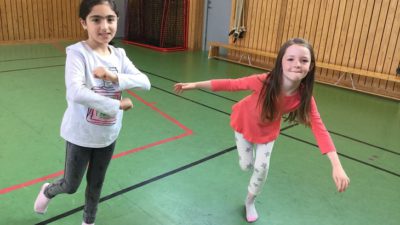 Judith och Kavi står i en gymnastiksal och dansar. En av dem snurrar armarna framför sig och den andra står på ett ben med armana utåt.