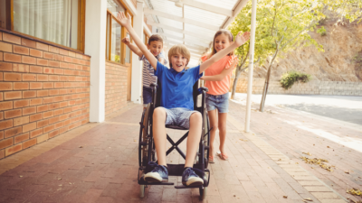 En pojke i blå tröja sitter i en rullstol. Han sträcker ut armarna och ser glad ut. Bakom honom står två andra personer och puttar på.