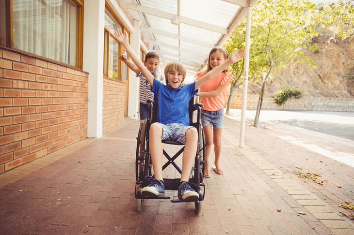 En pojke i blå tröja sitter i en rullstol. Han sträcker ut armarna och ser glad ut. Bakom honom står två andra personer och puttar på.