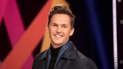 David står framför Melodifestivalens logga. Han har håret bakåtkammat och har på sig en grå jacka. Han ler stort mot kameran.