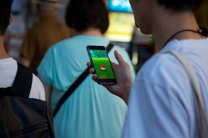 Ryggar på flera människor. Den som står närmast har en mobil i handen. På skärmens syns Pokémon Go-spelet.