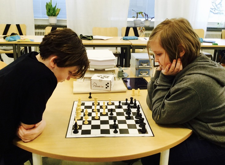 Två barn sitter mitt emot varandra i ett klassrum och tittar på ett schackbräde som ligger på bordet.