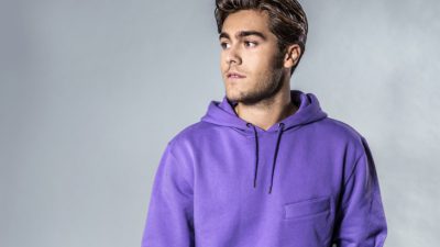 Benjamin framför en grå vägg. Han har en lila hoddie och halvkort, blont hår som ligger lite slarvigt bakåt. Han har lite skäggstubb och tittar åt sidan.