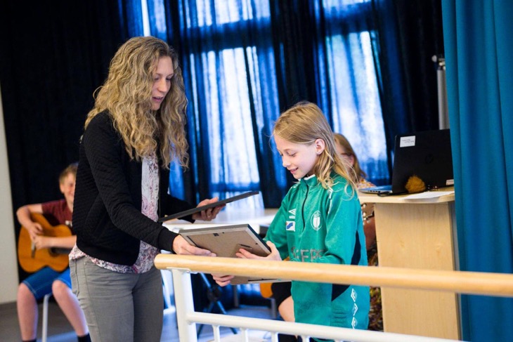 En vuxen håller i två inramade diplom. Barnet tar emot det ena. De är i ett stort rum med stora fönster med fördragna blåa gardiner.