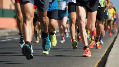 Många ben som springer. De flesta har färgglada springskor på sig. De springer på asfalt och solen skiner.