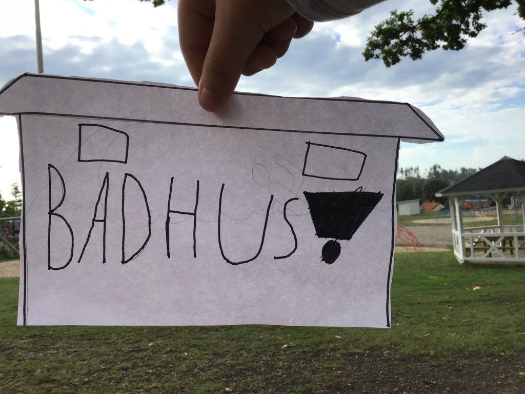 En ritad bild framför ett landskap. På bilden står det "badhus".