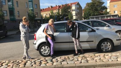 Tre barn håller för munnen vid en bil på en parkering.