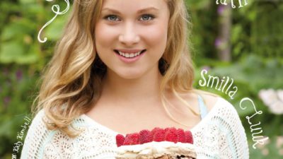 En bild på boken. Smilla står utomhus på sommaren. Hon ler och håller i en tårta som består av flera lager maräng med hela hallon och grädde emellan.