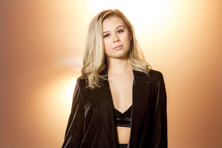 Lisa tittar rakt in i kameran med ett neutralt ansiksuttryck. På överkroppen har en kort topp på sig med en kavaj över. Hon har halvlångt hår och mycket smink.