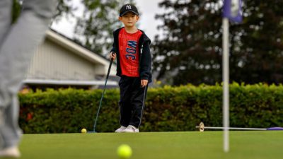 Erik står på golfbanan. Han har keps och träningsbyxor på sig. Han har klubben i handen. Han tittar bort mot bollen som han har puttat.
