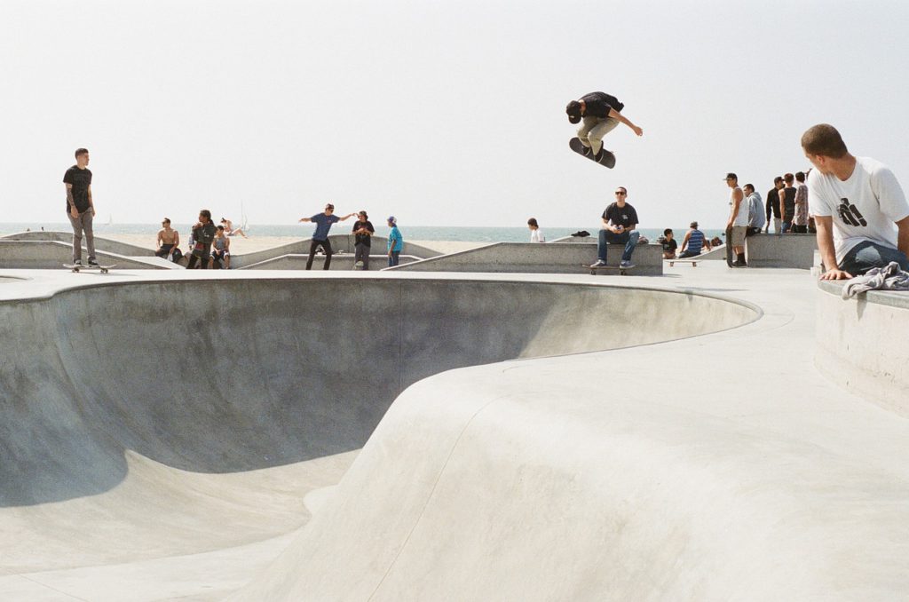 En skateboardpark vid havet. En person åker i en bowl och hoppar högt upp i luften med brädan. Runt om står människor och tittar på.