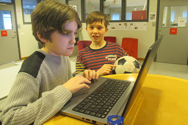 En kille sitter framför datorskärmen. Bredvid honom står en kompis med en fotboll.